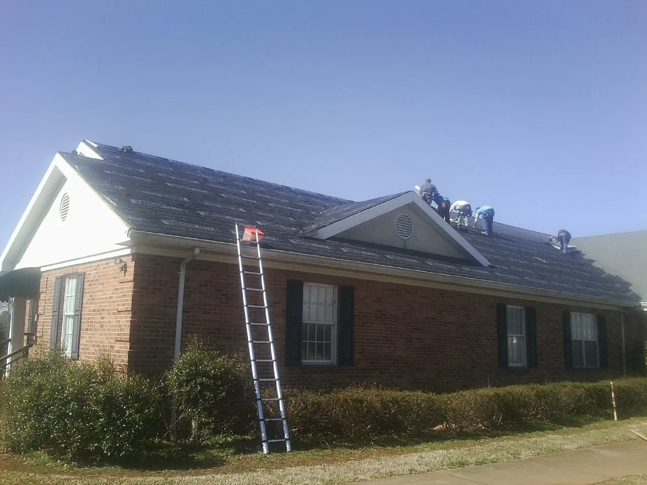 Crew tearing shingles off a brick ranch roof with a ladder set — Metal Roof Repair work by our crew in Walnut Grove