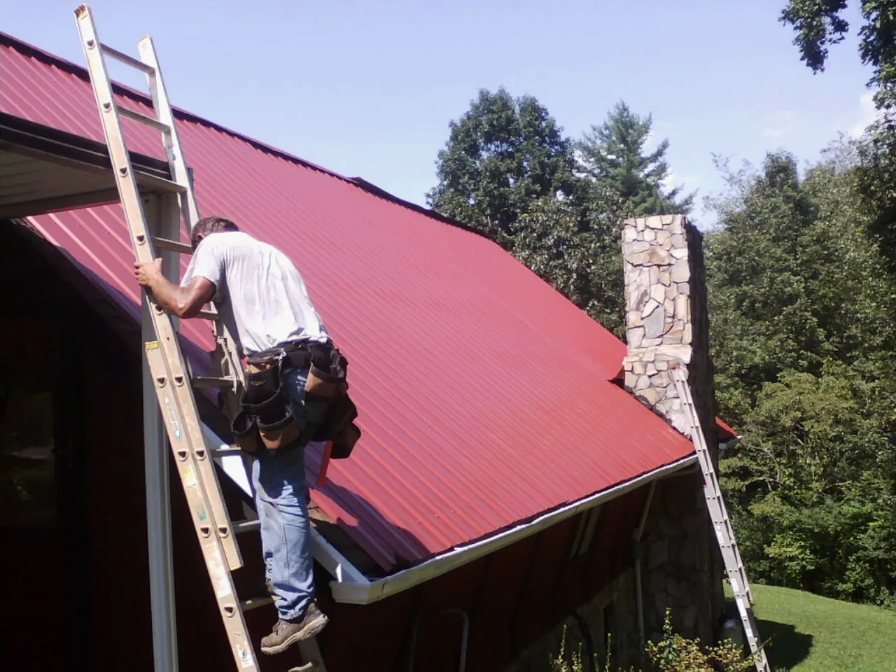 Roofer on a ladder beside a red metal roof and stone chimney — Soffit & Fascia Repair by Roofing Brothers in Walnut Grove
